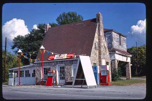 A vintage gas station with stone architecture, red pumps, and colorful banners under a blue sky with fluffy clouds.