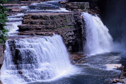 A scenic view of two waterfalls cascading over rocky cliffs into a river below, surrounded by lush greenery.