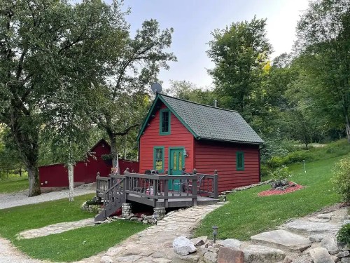 A charming red cabin with green trim, surrounded by trees and a stone pathway, set in a serene outdoor landscape.