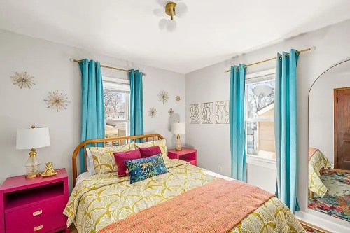 Bright bedroom featuring a bed with colorful bedding, pink nightstands, and turquoise curtains by sunny windows.