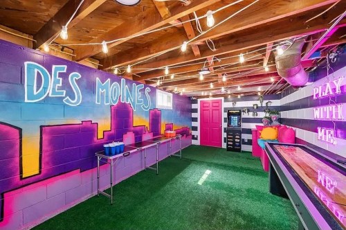 A colorful game room with a mural of Des Moines, bright lights, and playful decor, featuring a pink door and green turf.