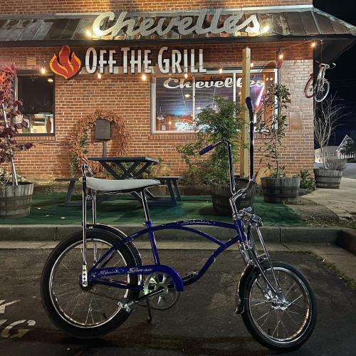 A blue bicycle parked in front of a restaurant named "Chevelle's Off the Grill" at night.