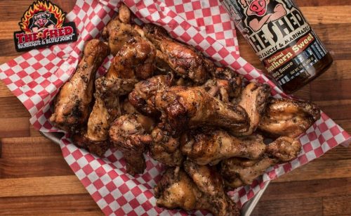 A platter of grilled chicken wings on red-checkered paper, alongside a bottle of barbecue sauce.
