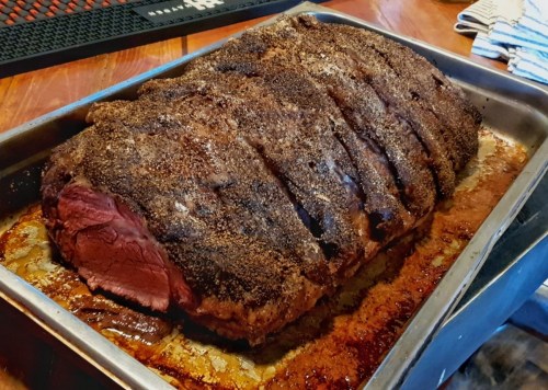 A large, seasoned roast beef resting in a metal tray, with a crispy crust and juicy interior, on a wooden table.