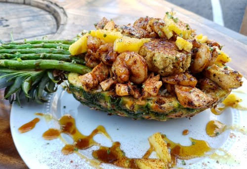 A halved pineapple filled with shrimp, chicken, and diced fruit, garnished with spices and drizzled with sauce.
