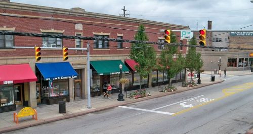 A street view of a small town with shops, colorful awnings, traffic lights, and trees lining the sidewalk.