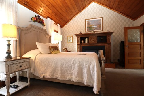 Cozy bedroom with a wooden ceiling, a bed with white linens, a nightstand, and a decorative fireplace.