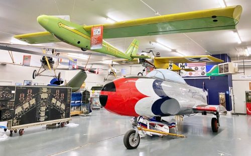 A green and yellow glider hangs above a red, white, and blue aircraft in a spacious aviation museum.