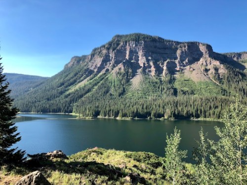 A serene lake surrounded by lush green forests and a towering mountain under a clear blue sky.