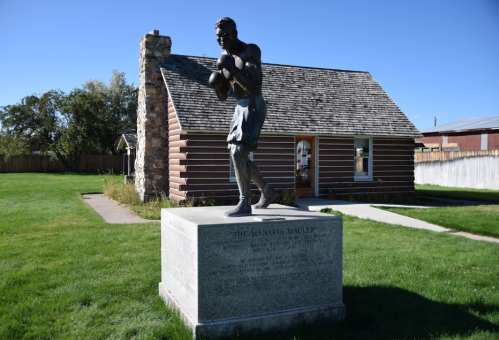 A bronze statue of a boxer stands in front of a log cabin, surrounded by green grass and clear blue skies.