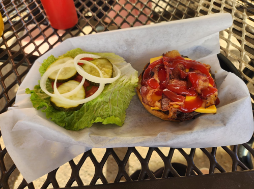 A burger with bacon, cheese, and ketchup next to a side of lettuce, pickles, and onions on a table.