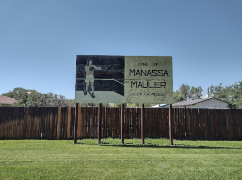 Sign reading "Home of Manassa Mauler Jack Dempsey" with a vintage illustration of a boxer, set against a clear blue sky.