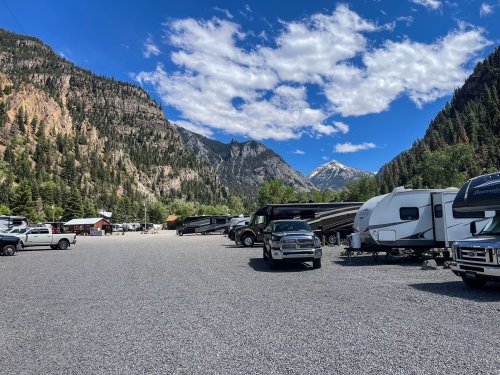 A gravel parking area filled with RVs and trucks, surrounded by mountains and a blue sky with scattered clouds.
