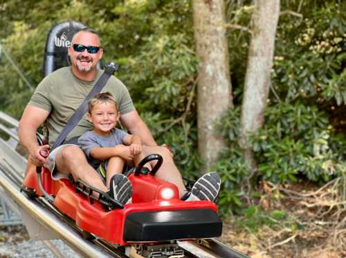 A man and a boy smile while riding a red alpine coaster through a wooded area.