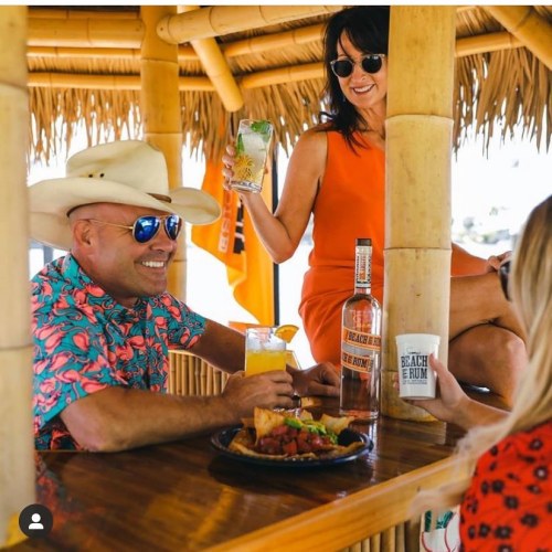 A man in a cowboy hat and a woman in a red dress enjoy drinks at a beach bar with food on the table.