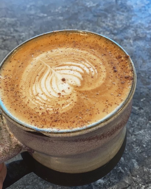 A close-up of a latte in a textured mug, featuring intricate foam art and a sprinkle of cocoa on top.