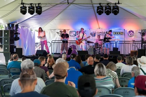 A band performs on stage under a tent, with an audience seated in front, enjoying the live music.