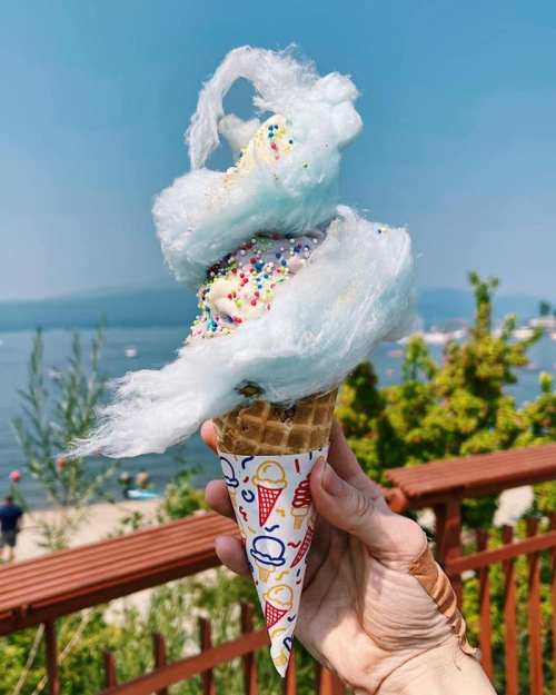 A hand holds an ice cream cone topped with blue cotton candy and colorful sprinkles, with a scenic background.