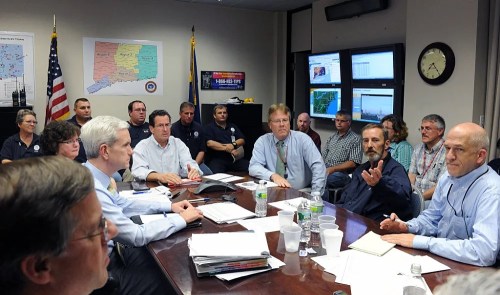 A group of people in a conference room discussing plans, with maps and screens visible in the background.