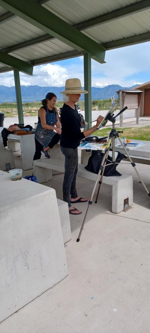 A woman in a wide-brimmed hat paints on an easel while another woman sits nearby in a shaded outdoor area.