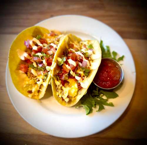 Two tacos filled with meat, topped with diced tomatoes and lettuce, served with a side of salsa on a plate.