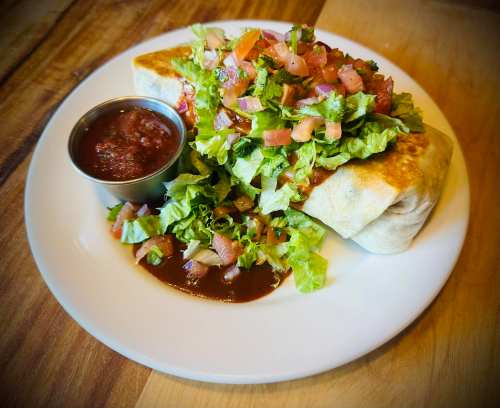 A burrito topped with fresh lettuce and tomatoes, served with a side of salsa on a white plate.