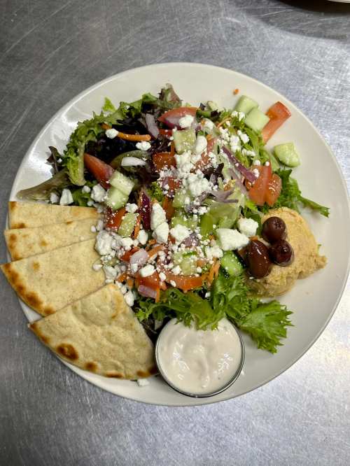 A fresh salad with mixed greens, tomatoes, cucumbers, olives, feta cheese, pita bread, and a side of dressing.