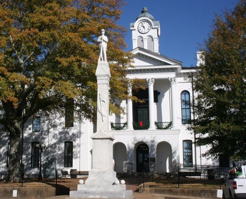 A white monument of a soldier stands in front of a historic building with a clock tower and autumn trees.