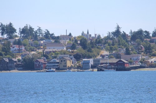 A coastal view of a small town with colorful buildings and trees, set against a clear blue sky and calm water.