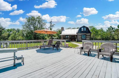 A spacious deck with chairs and an umbrella, overlooking a log cabin and lush greenery under a blue sky with clouds.