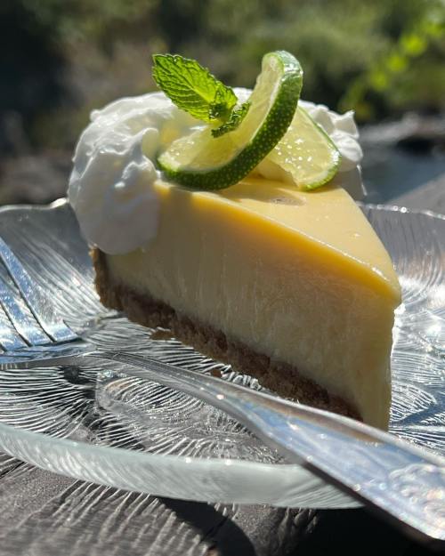 A slice of lime cheesecake topped with whipped cream and lime slices, served on a glass plate with forks.