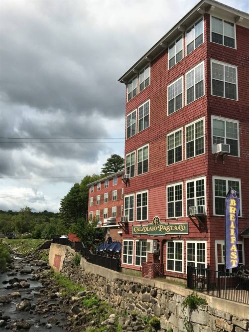 A red-brick building with large windows beside a rocky stream, featuring a sign for a pasta restaurant and breakfast.