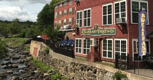A red brick building with a sign for "Capriolo Pasta Co" beside a stream and outdoor seating area.