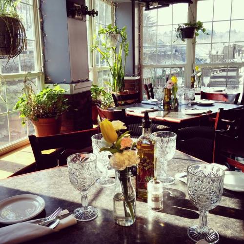 Brightly lit restaurant interior with plants, elegant table settings, and a vase of flowers on a table.