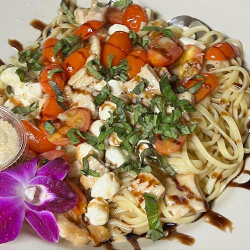 A plate of pasta topped with chicken, cherry tomatoes, basil, mozzarella, and a flower garnish, drizzled with balsamic.