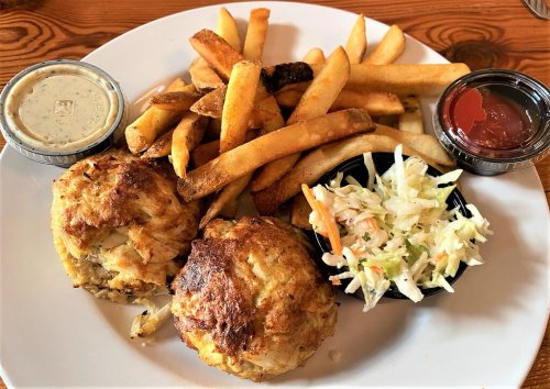 A plate with two crab cakes, French fries, coleslaw, and dipping sauces.