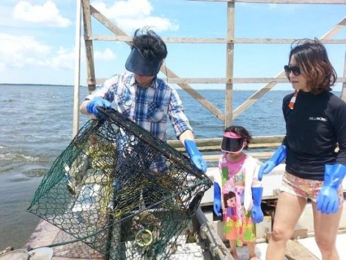 A man and woman, wearing gloves and visors, examine a crab trap with a young girl on a boat by the water.