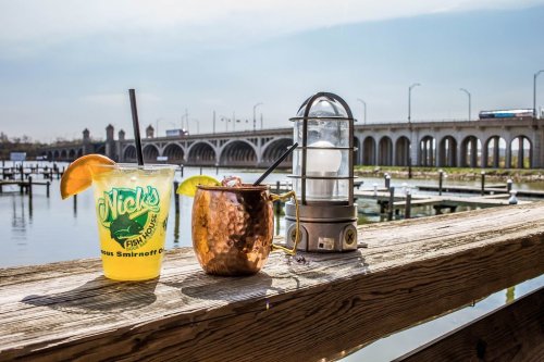 A vibrant cocktail and a copper mug sit on a wooden railing by a river, with a bridge and clear sky in the background.