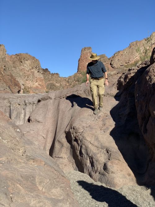 A person in a hat walks along a rocky terrain with steep cliffs under a clear blue sky.