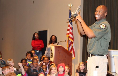 A smiling presenter holds an alligator while engaging a captivated audience of children in a classroom setting.