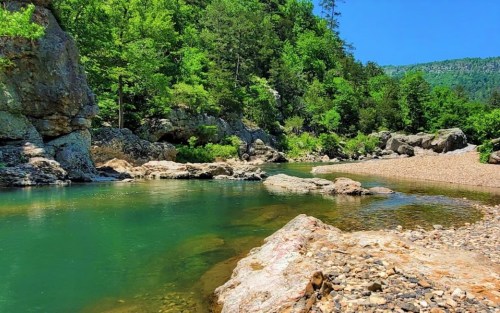 A serene river flows through a lush green landscape, surrounded by rocky banks and trees under a clear blue sky.