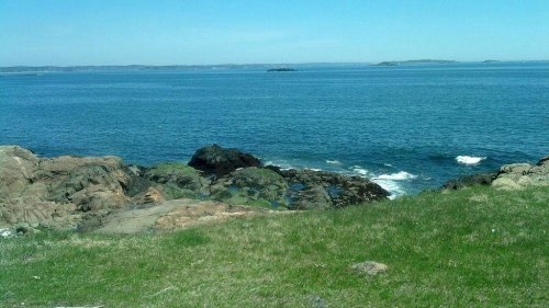 A serene coastal view featuring rocky shores, gentle waves, and a clear blue sky over the water.