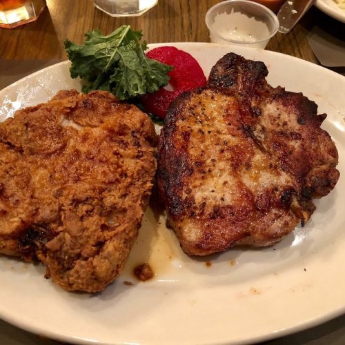 A plate featuring a crispy fried chicken breast, a grilled pork chop, and a side of greens and tomato.