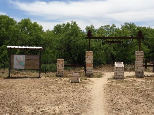 Entrance to a park with stone pillars, a wooden sign, and informational boards surrounded by greenery and a dirt path.