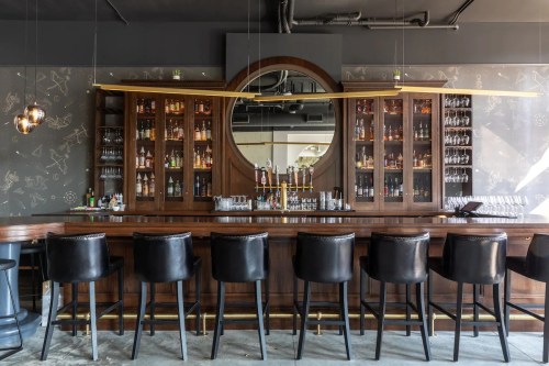 A stylish bar with a wooden counter, black stools, and shelves filled with bottles and glassware against a patterned wall.