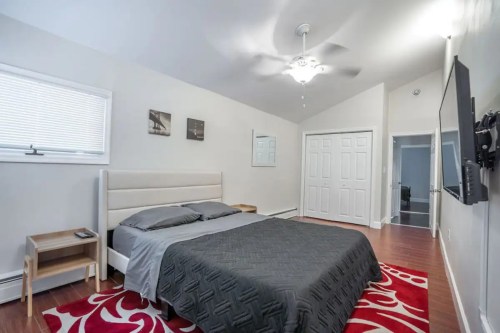 A modern bedroom featuring a queen bed, nightstands, a ceiling fan, and a large TV on the wall. Red area rug on the floor.