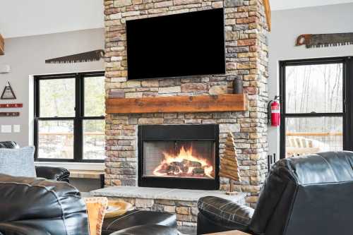 Cozy living room with a stone fireplace, black TV, and leather chairs, surrounded by large windows and winter scenery.