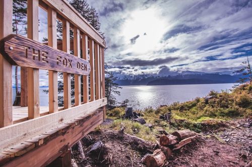 A wooden sign reading "The Fox Den" overlooks a serene lake and mountains under a cloudy sky.
