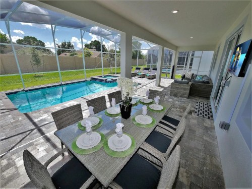 A modern patio with a dining table set for eight, overlooking a pool and lush green lawn through a screened enclosure.