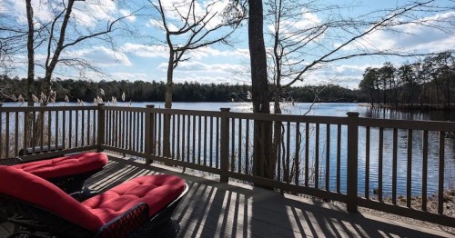 A serene lakeside view from a deck with red lounge chairs, surrounded by trees and a clear blue sky.